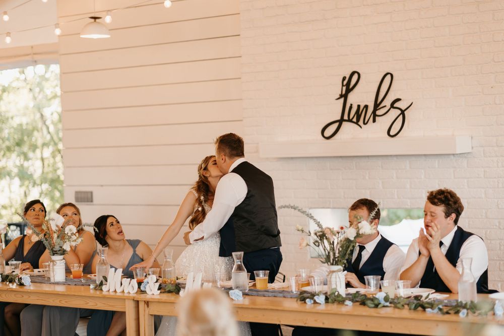 Bride and groom share a kiss at the head table during their wedding reception at a rustic barn venue.