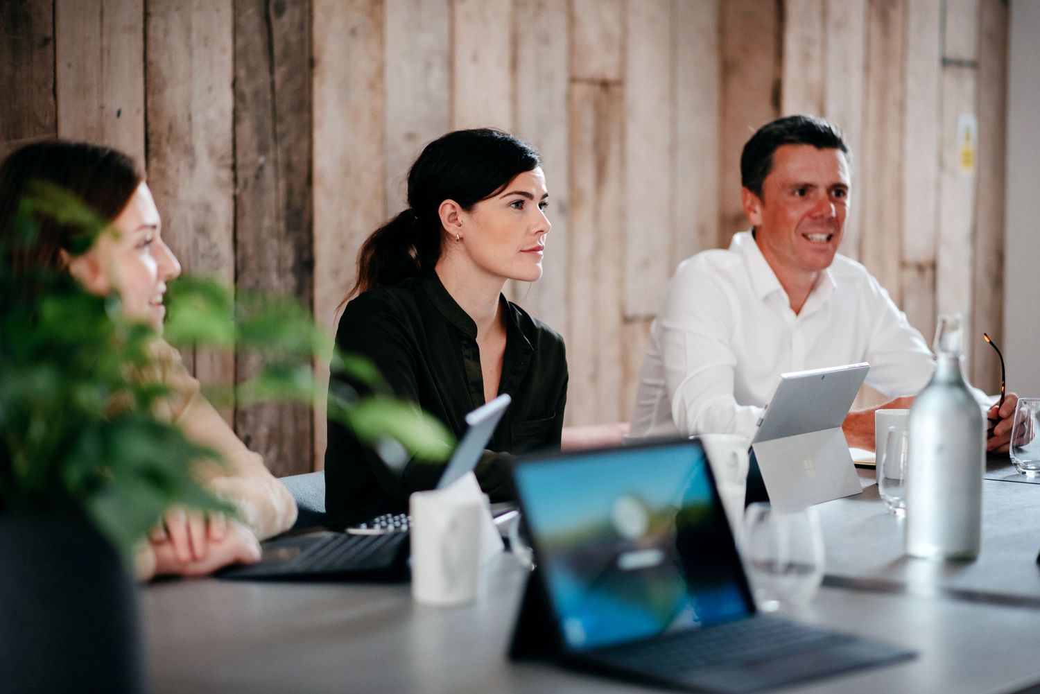Three people meet around a conference table with laptops and water bottles in a business setting.