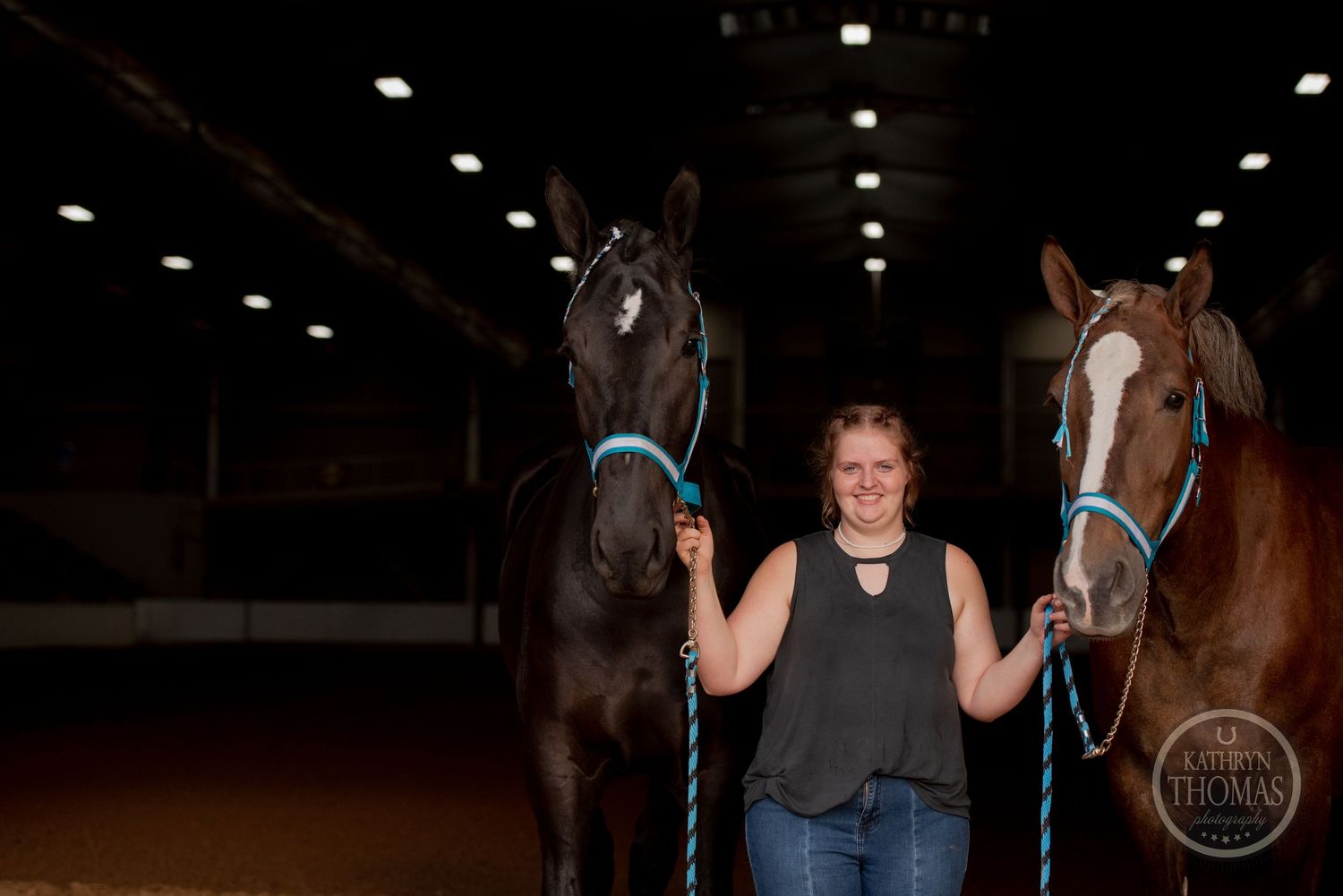 Two striking horses with blue lead ropes pose in a dimly lit indoor arena.