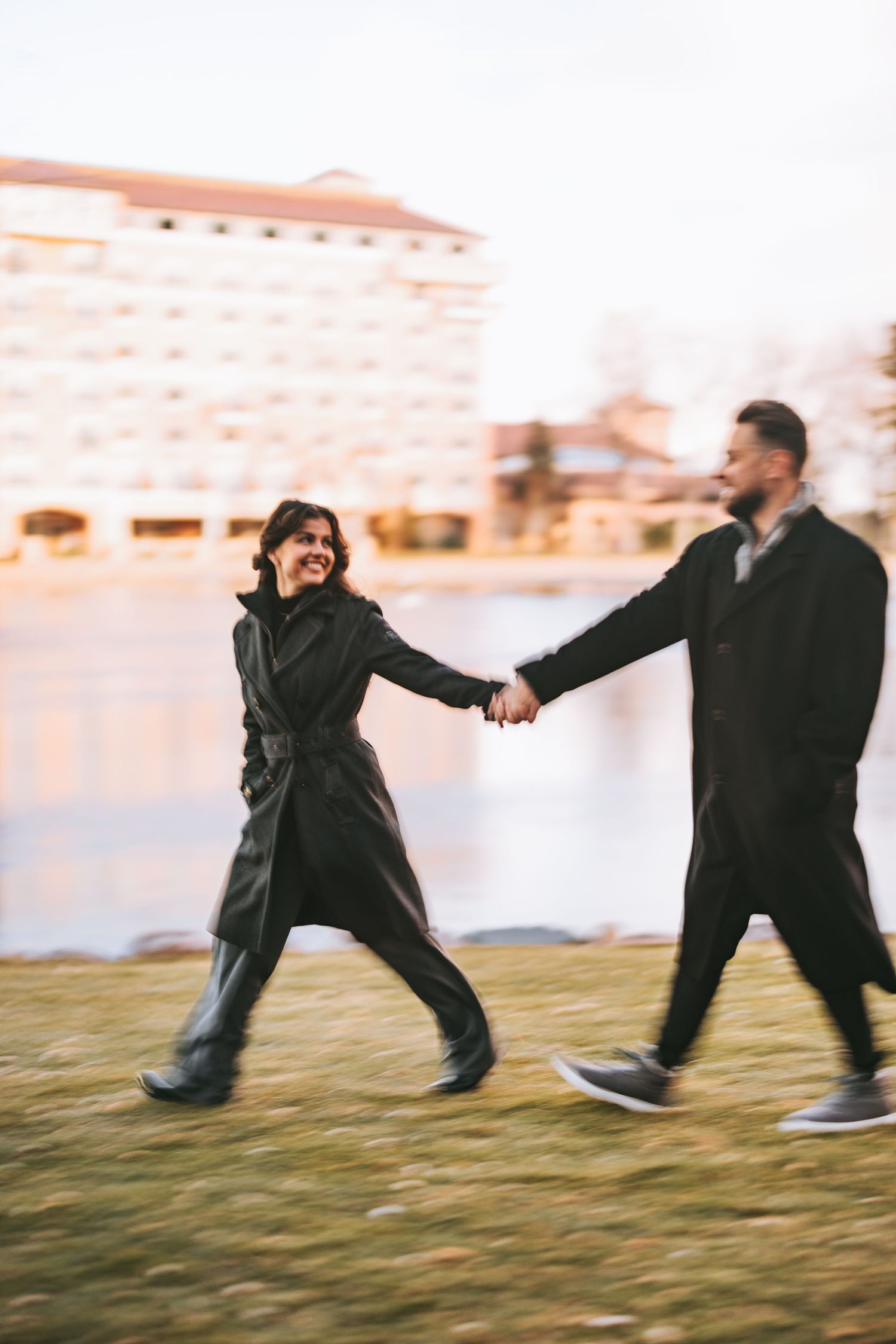A couple playfully running and holding hands while wearing black winter coats on grass.