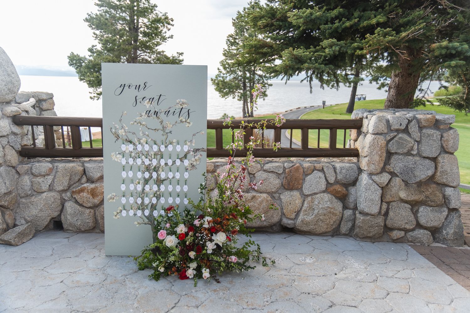 A seating chart display with wine glasses arranged on an elegant white board next to a stone wall overlooking the water.