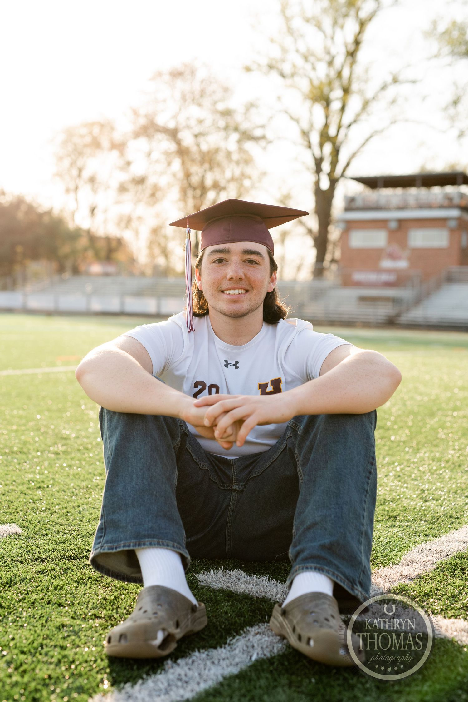 Graduate in cap and gown sits relaxed on athletic field during sunset portrait session.