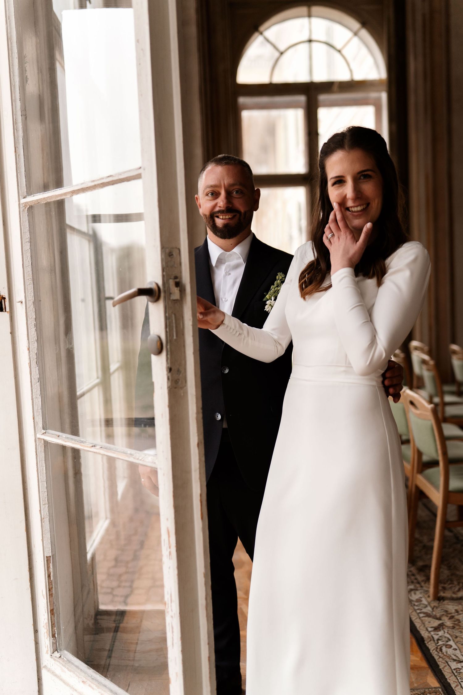 Elegant couple in wedding attire posing by vintage doorway with arched window.