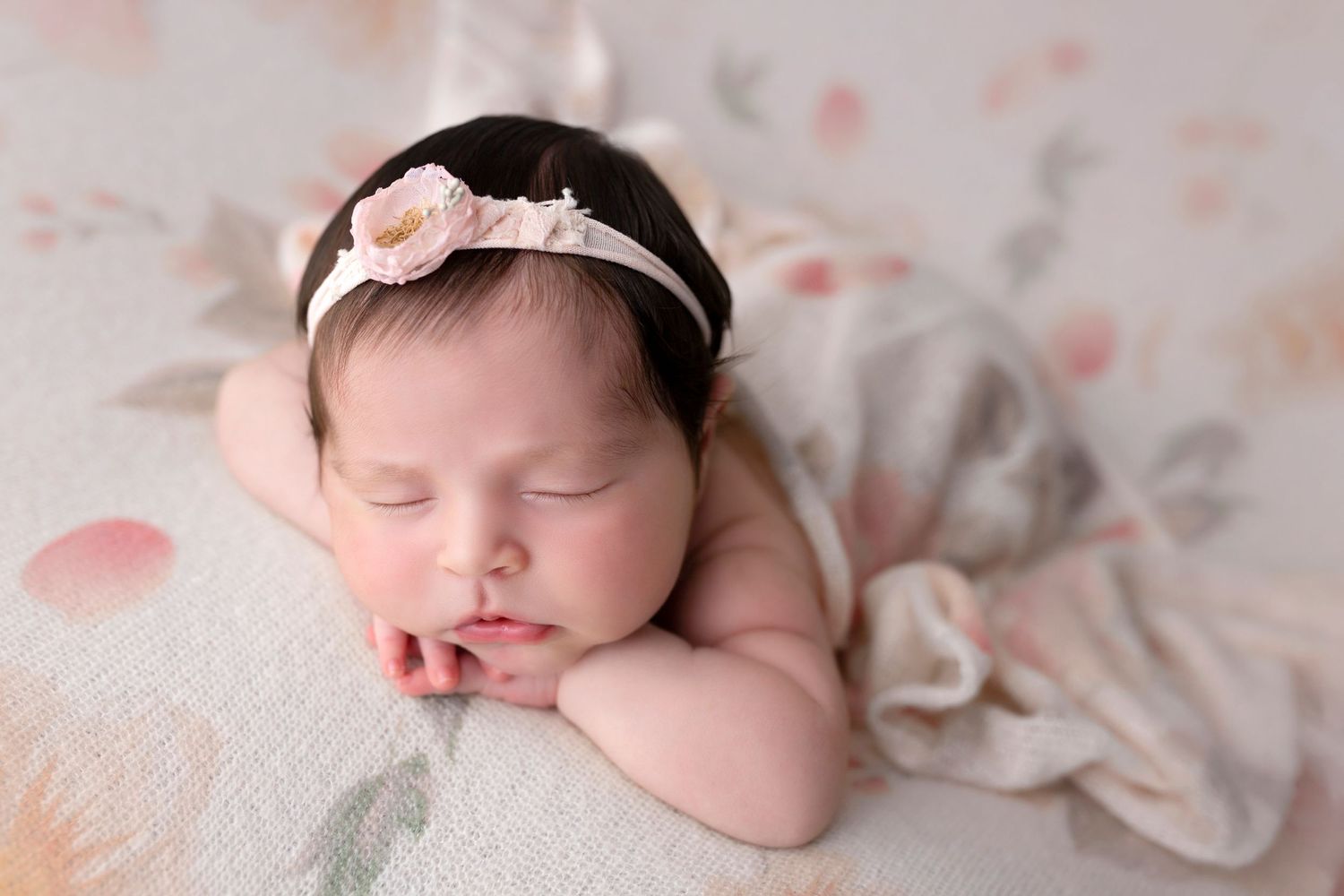 Sleeping newborn wearing floral headband on light textured backdrop.