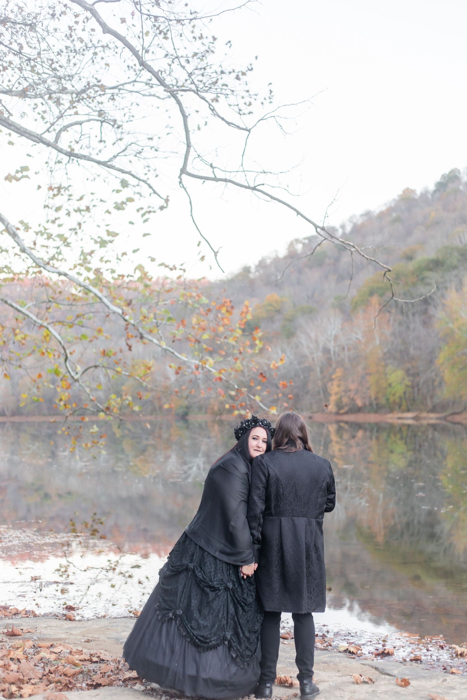 Two figures in dark clothing stand by a misty lake surrounded by autumn foliage.