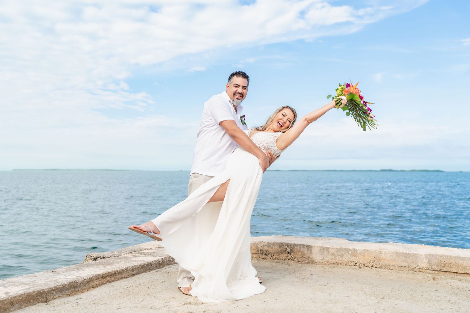 A joyful wedding couple poses playfully on a waterfront ledge against a blue ocean backdrop.