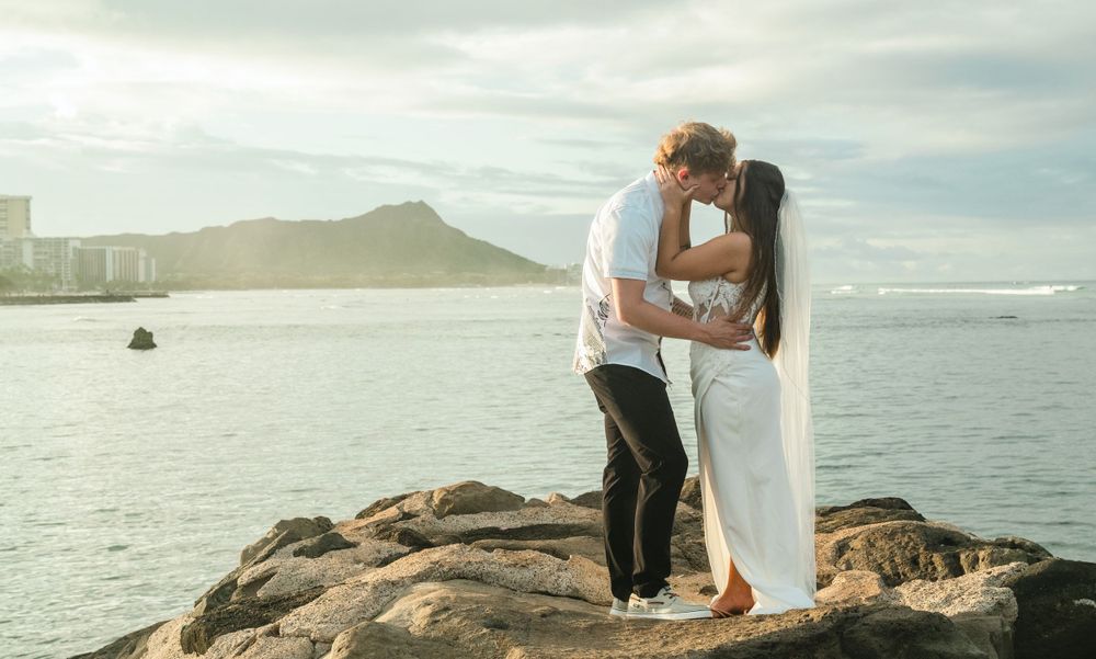 A tender moment between newlyweds standing on oceanside rocks with Diamond Head in background.