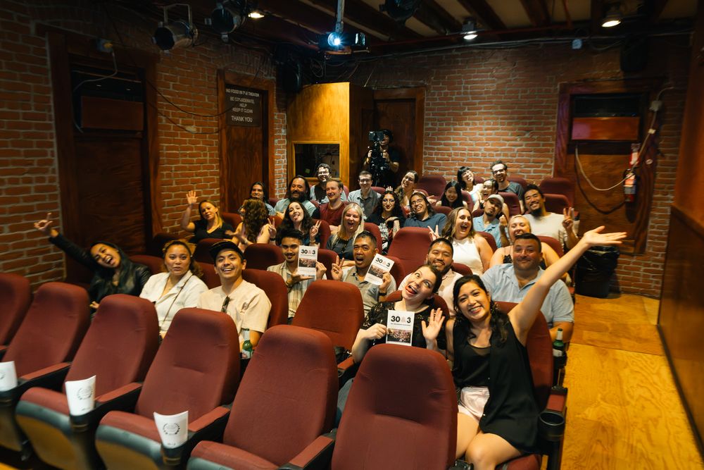Theater audience seated in burgundy chairs watching a performance in brick-walled venue.