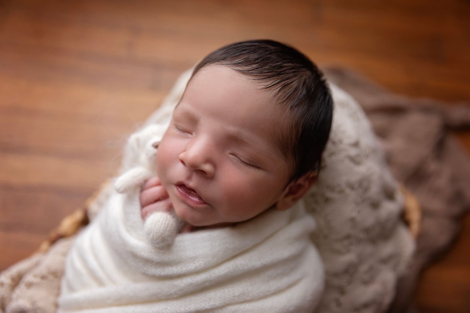 Sleeping newborn wrapped in cozy white swaddle on warm brown wooden surface.