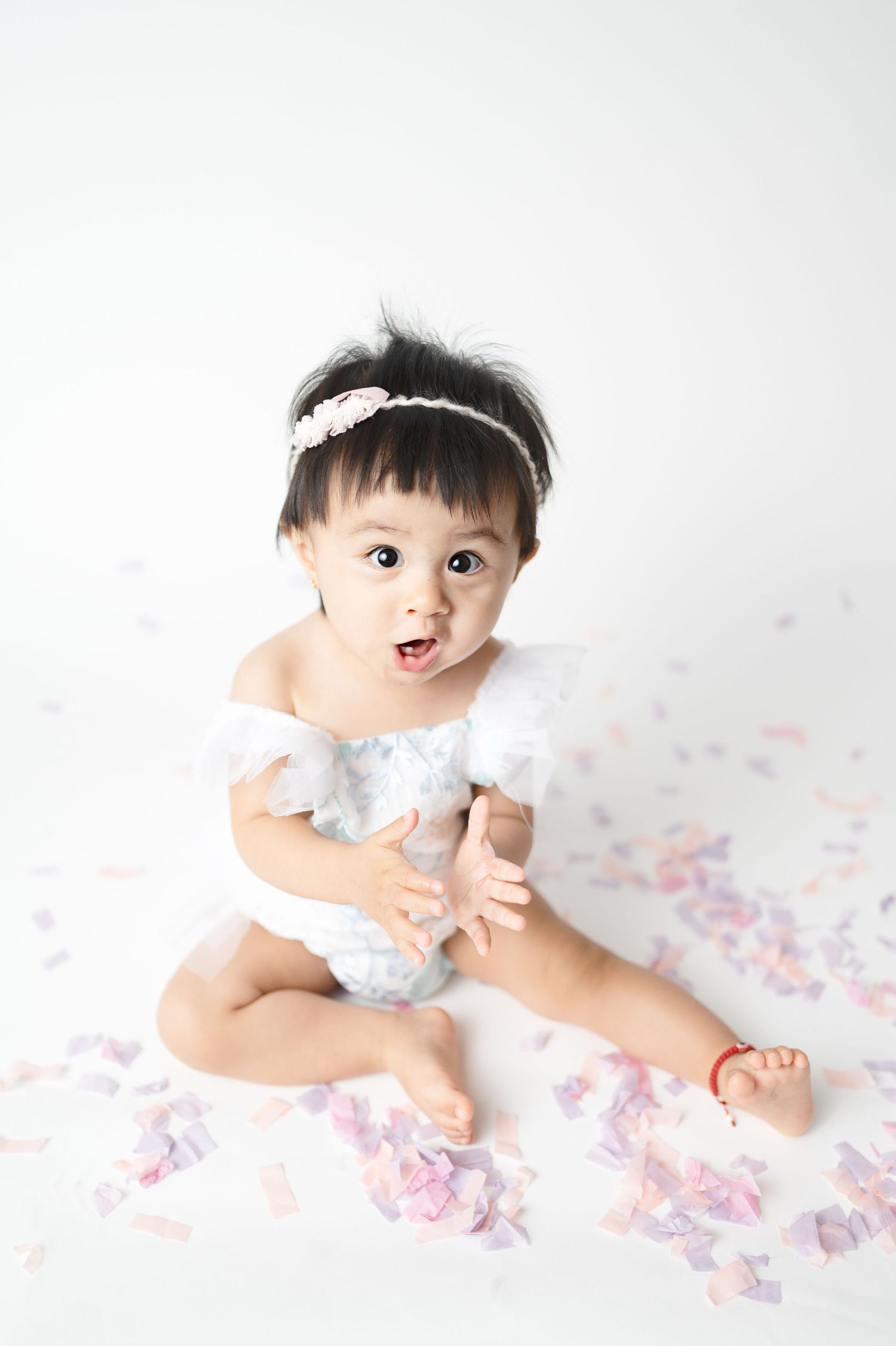 Baby in white outfit smiling and sitting among scattered pink petals.