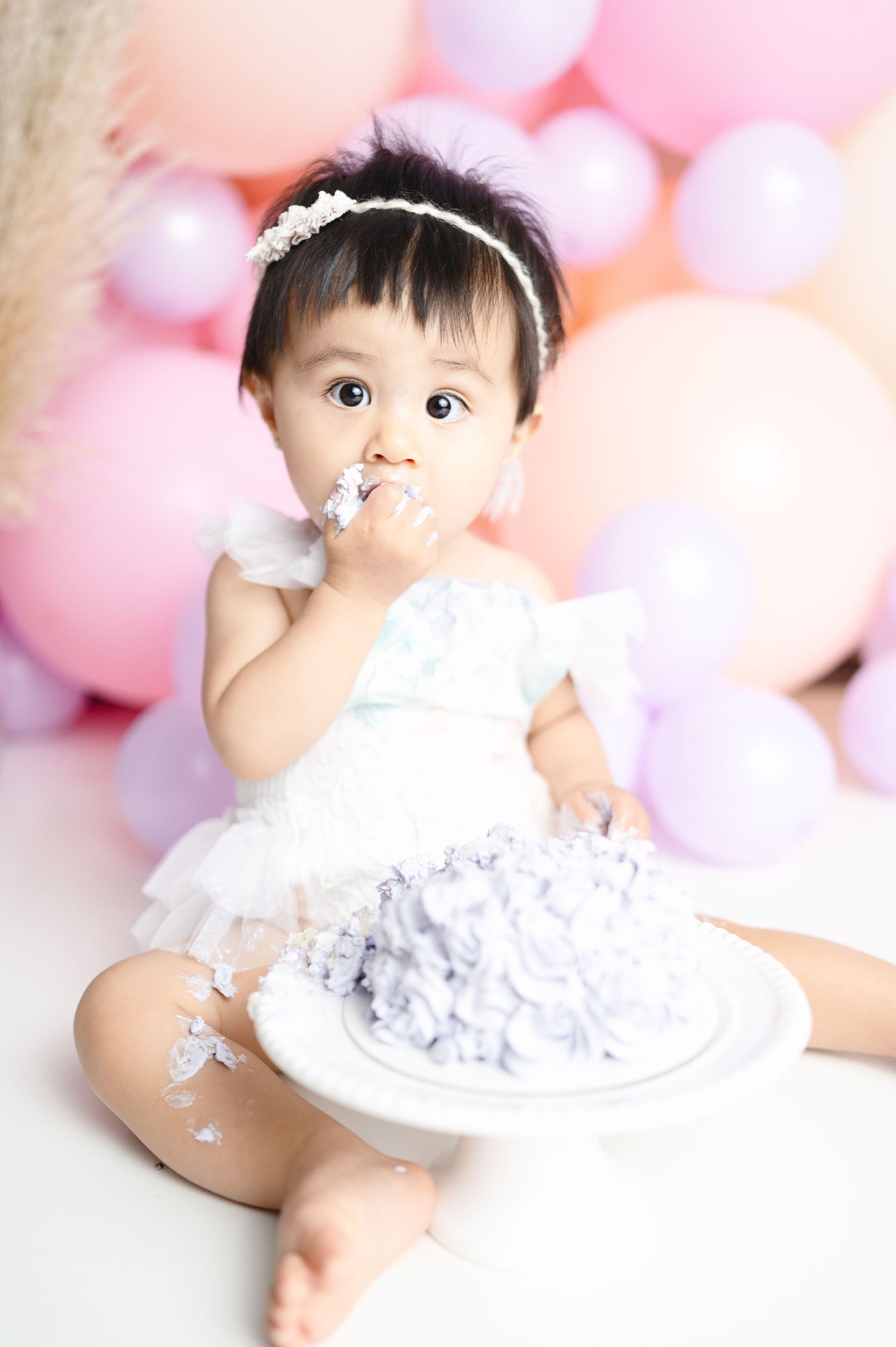 Baby in white dress eating cake during first birthday photoshoot with pink balloons.