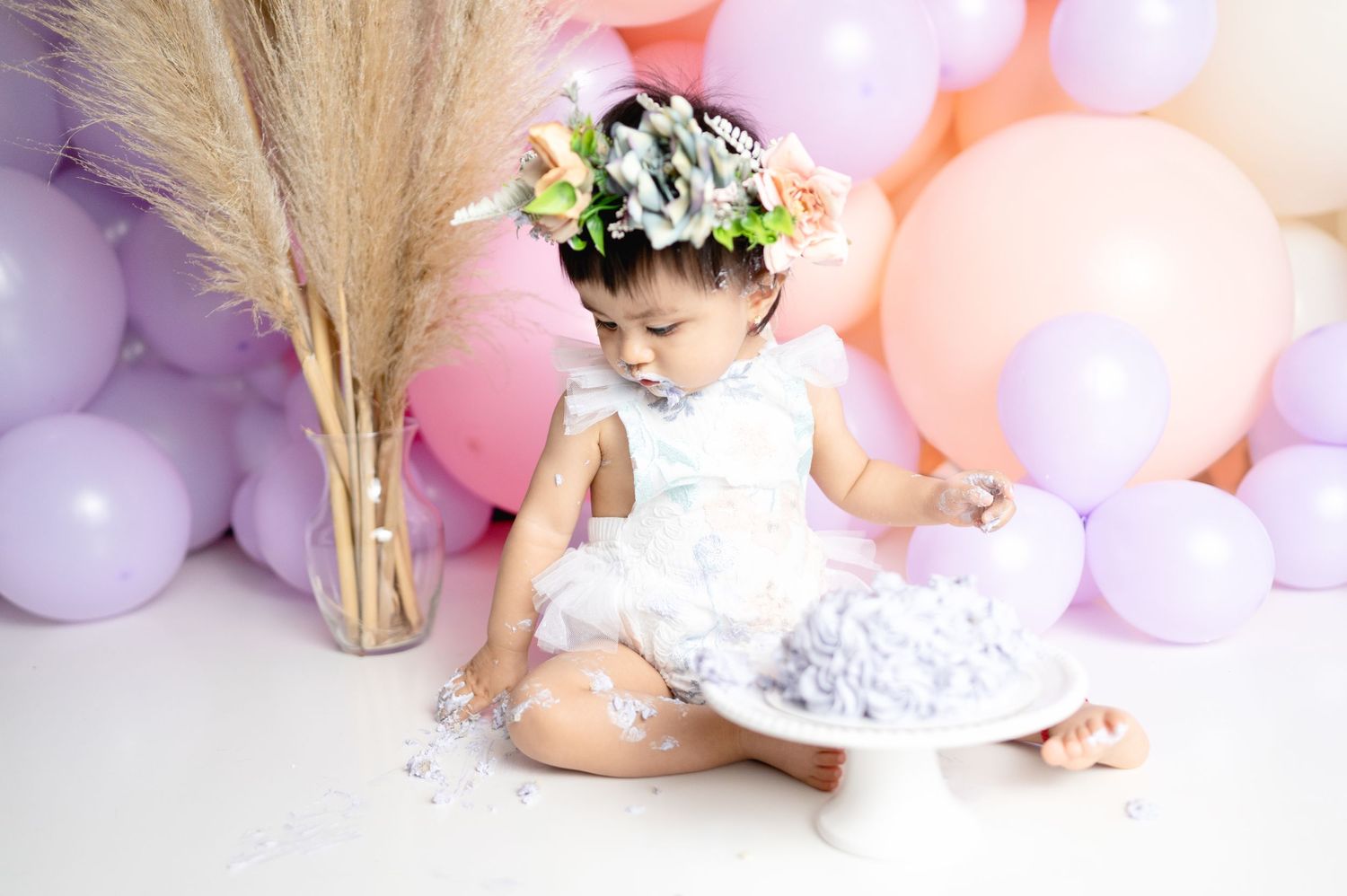 Baby in white dress with floral crown exploring cake with pink balloon backdrop.