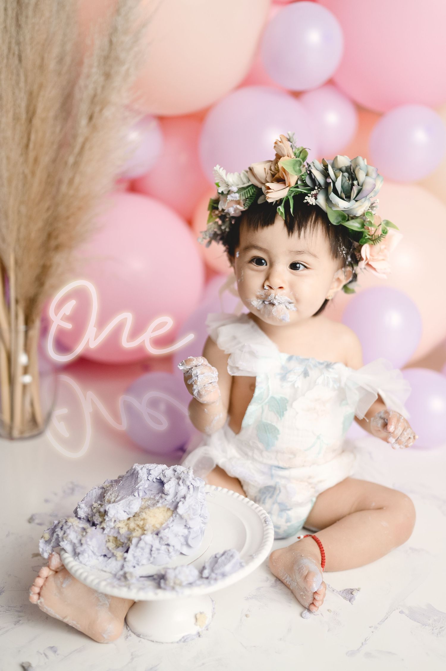 Baby with cake-covered face during first birthday photoshoot with neon 'One' sign.