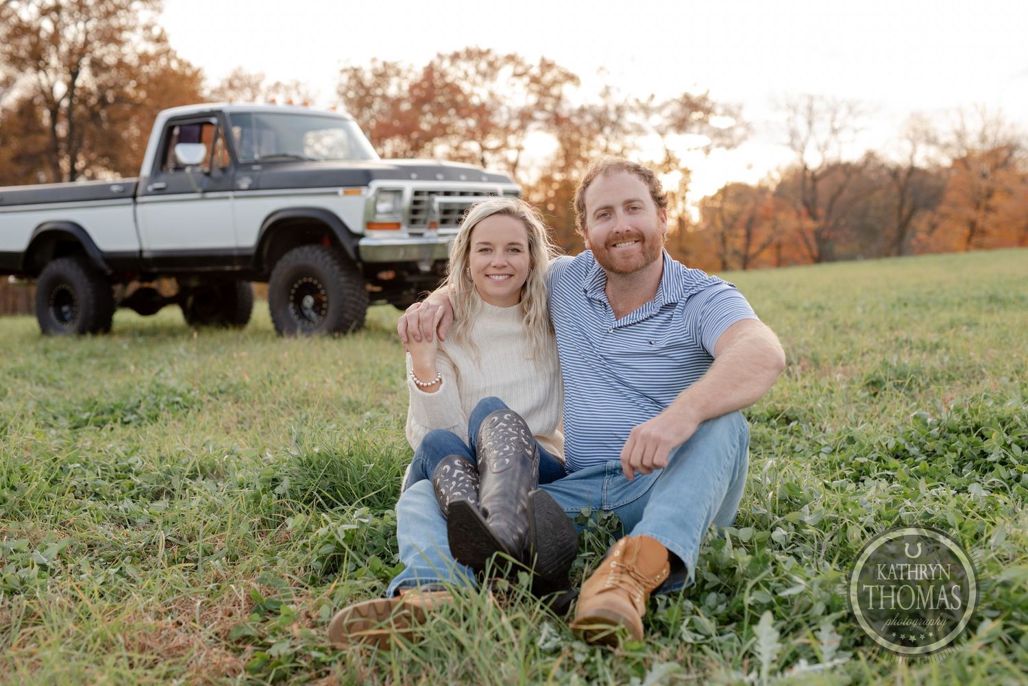 A couple sits together in a grassy field in front of a classic lifted Ford truck during autumn.