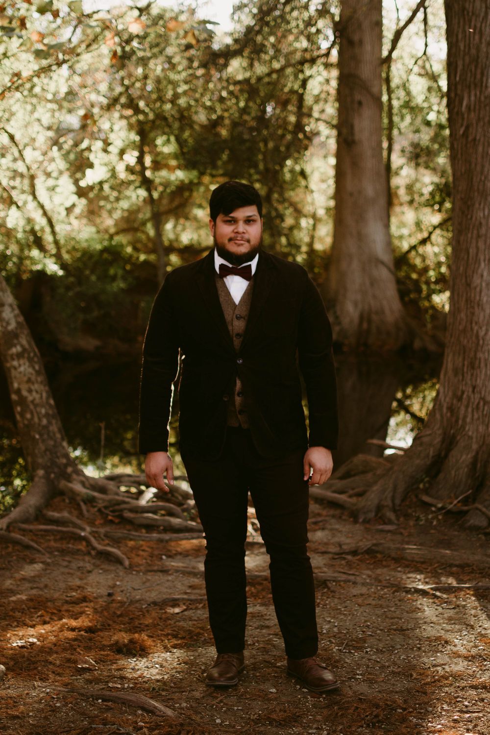 Formal portrait of person in classic tuxedo with brown vest standing in wooded setting.