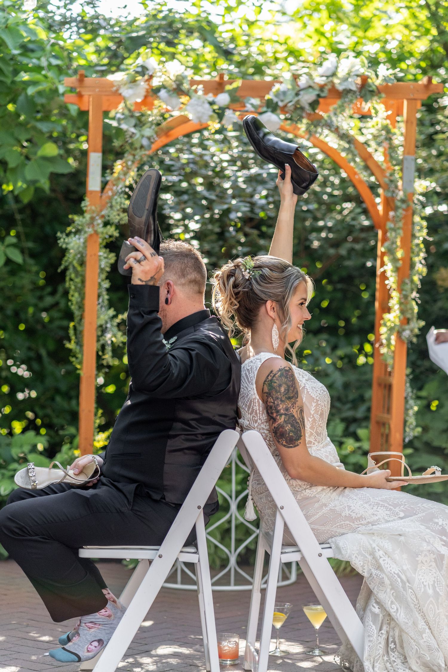 Playful bride and groom sitting back-to-back during wedding reception game under floral arch.
