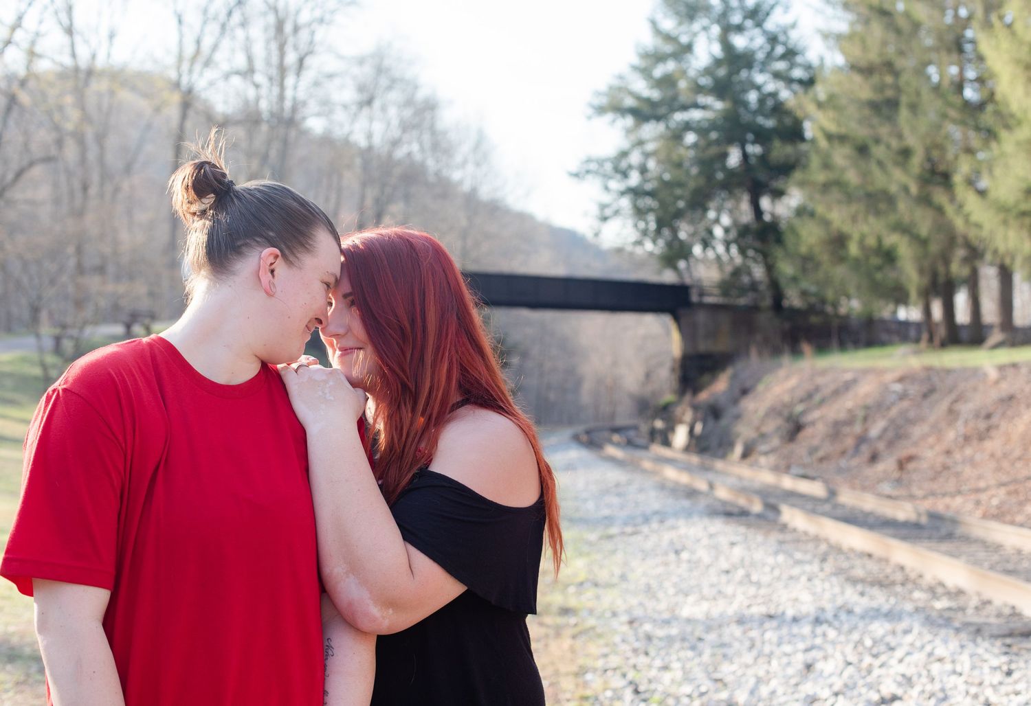 A couple shares a tender moment outdoors with a bridge visible in the background.
