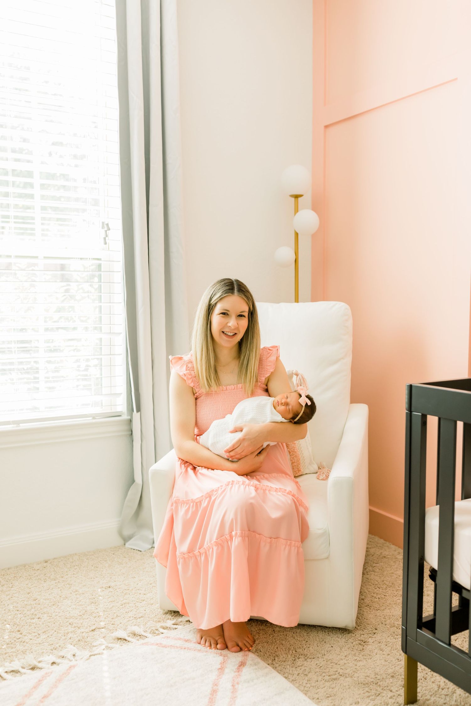 Woman in coral dress sits in white chair holding newborn baby in nursery.