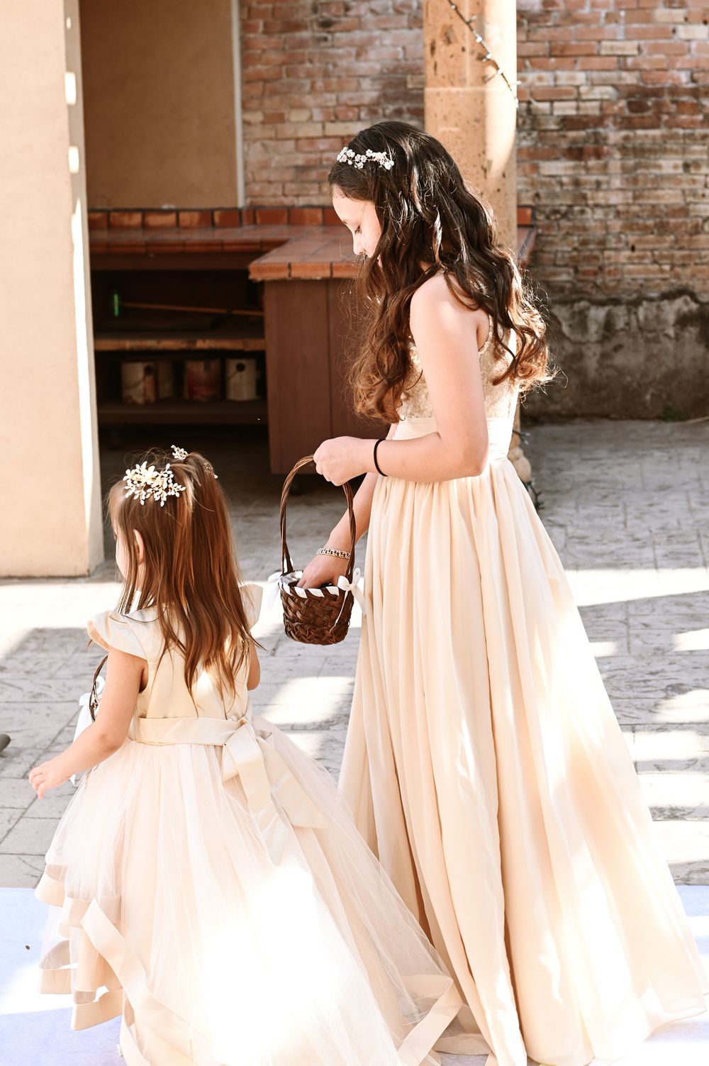 Two flower girls in cream-colored dresses carrying baskets in a sunlit brick corridor.