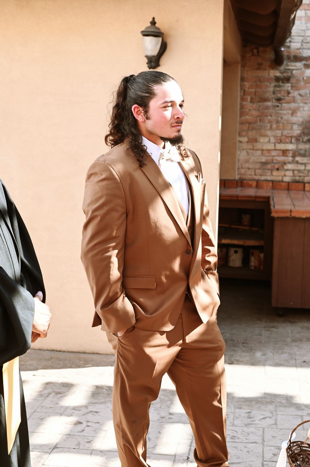 A person in a brown suit stands confidently in front of a brick wall and stucco exterior.