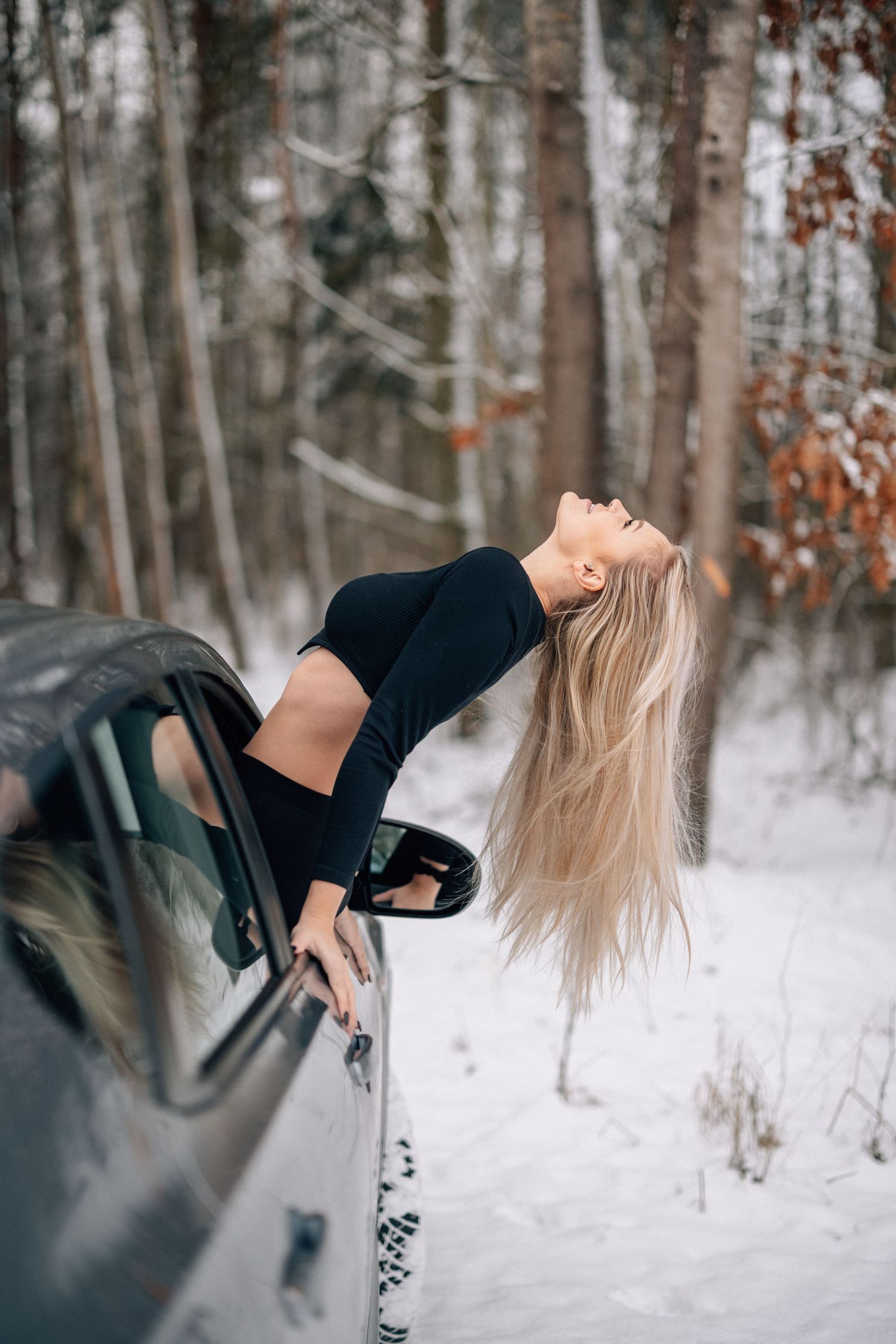 A creative portrait of someone leaning out of a car window in a snowy forest setting.