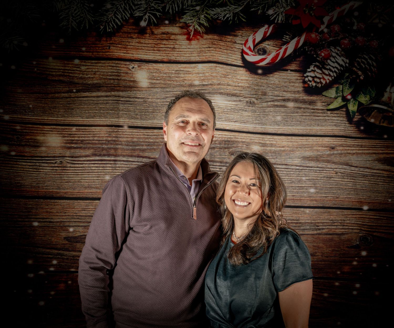 Couple posing together against wooden wall with holiday decorations and pine cones.