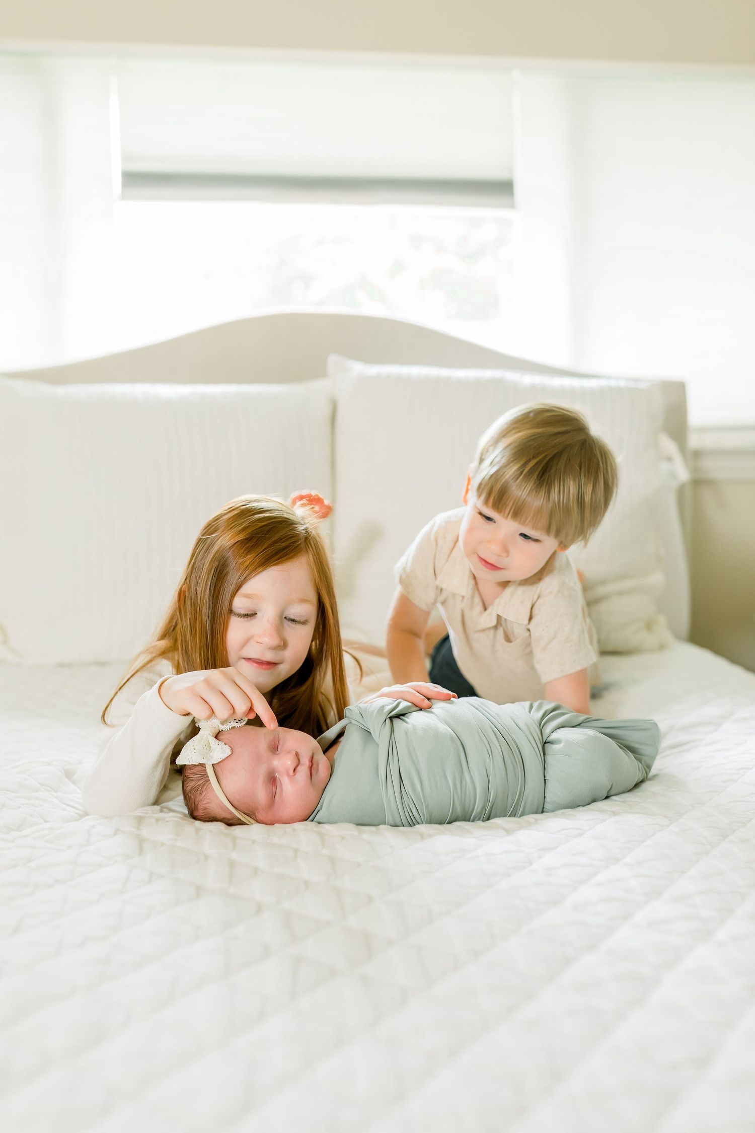 Two children lean over and interact with a swaddled baby on a white bed.