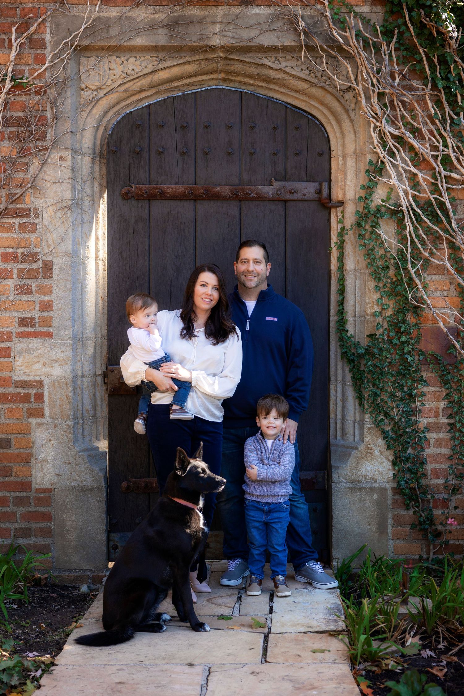Family with two small children and black dog pose in front of rustic wooden doorway with stone arch and brick walls.