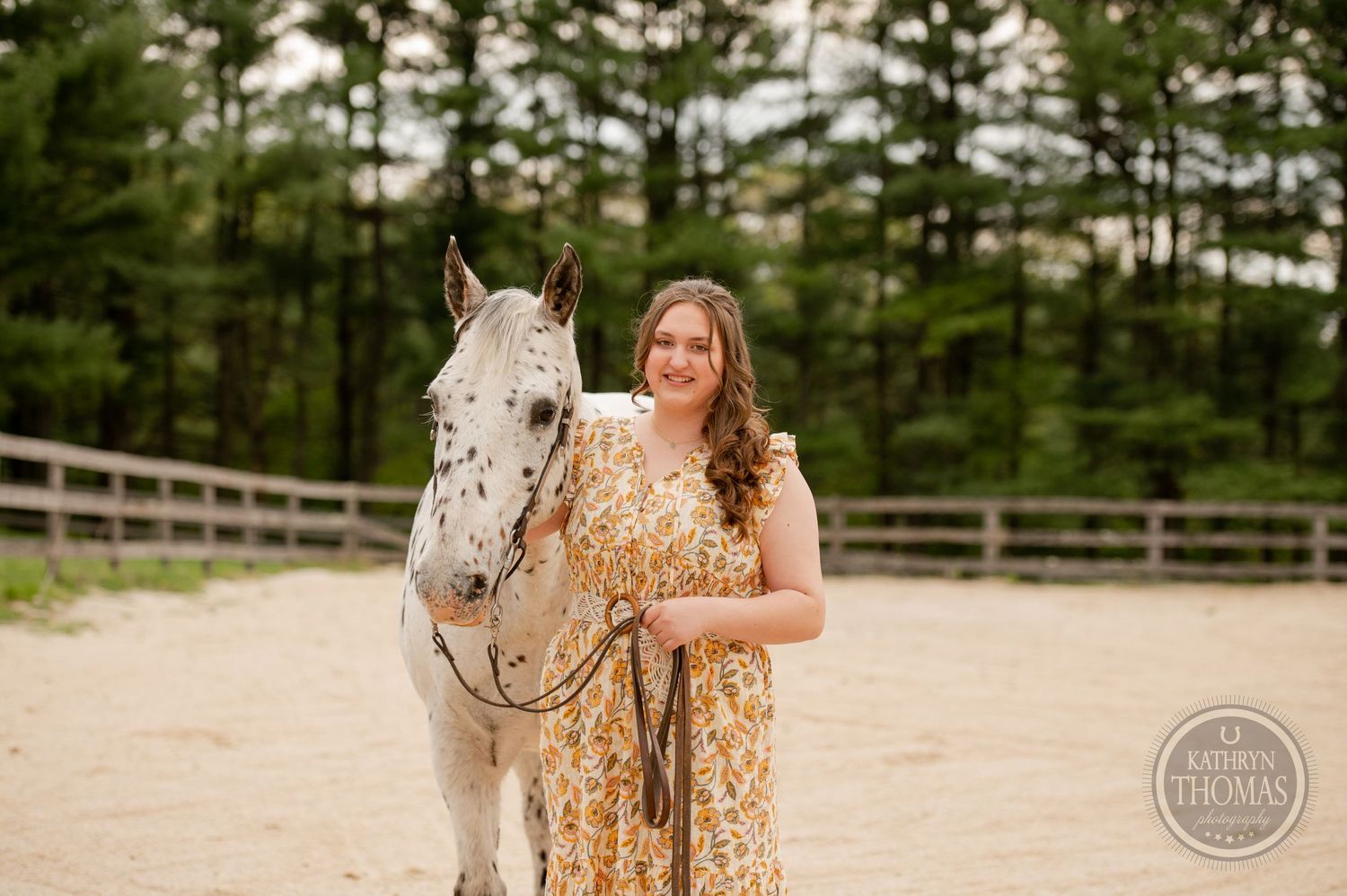 A spotted white horse and companion pose together in an enclosed arena with pine trees in the background.