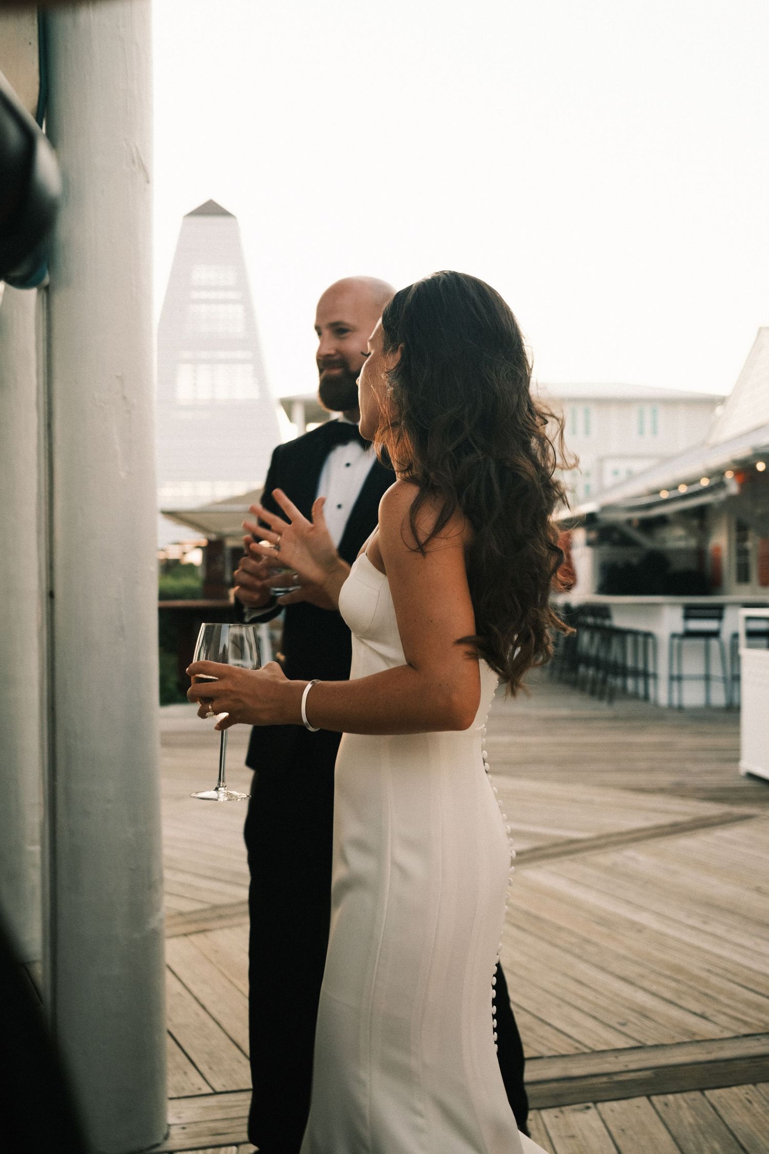 Couple in formal attire standing between white pillars on a waterfront deck.
