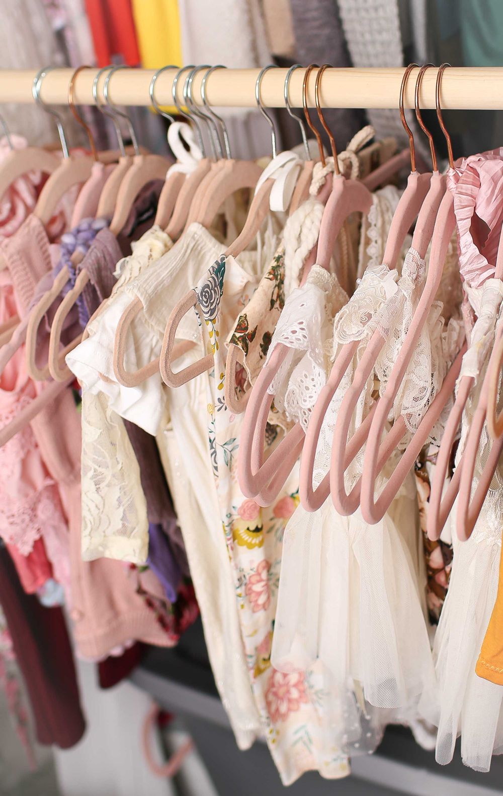 Pink and white baby dresses with floral patterns and lace details hang neatly on wooden hangers in a clothing rack.