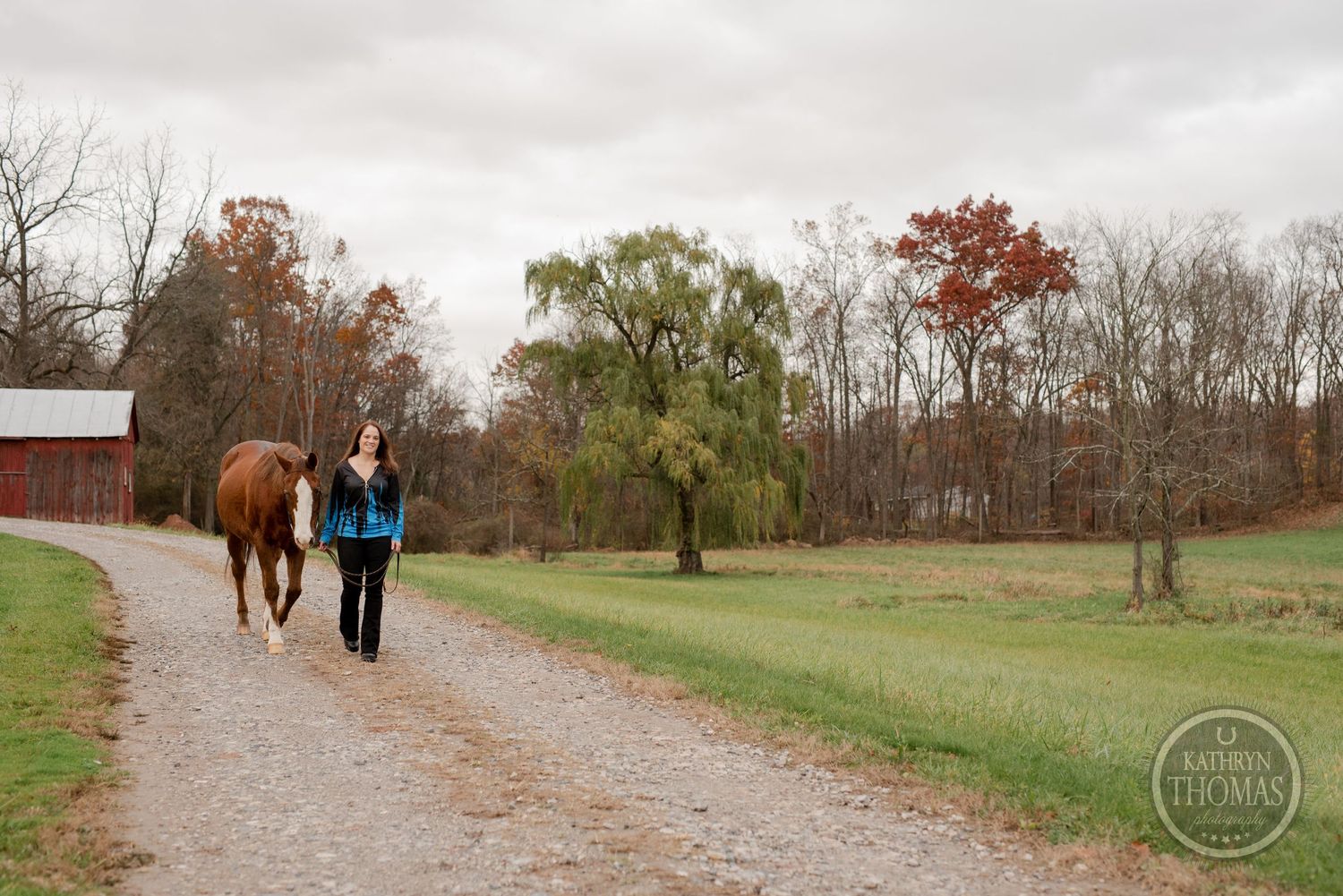 A chestnut horse and person walk along a country path near a red barn on an autumn day.