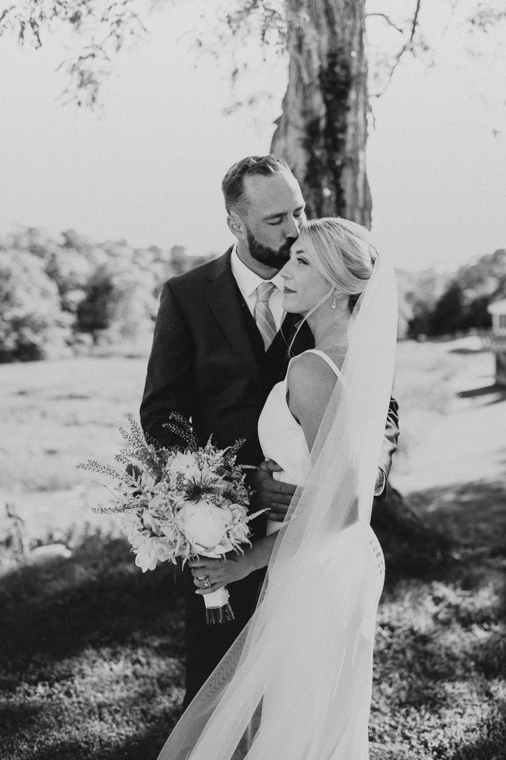 Black and white wedding portrait showing a tender moment between newlyweds holding a floral bouquet.