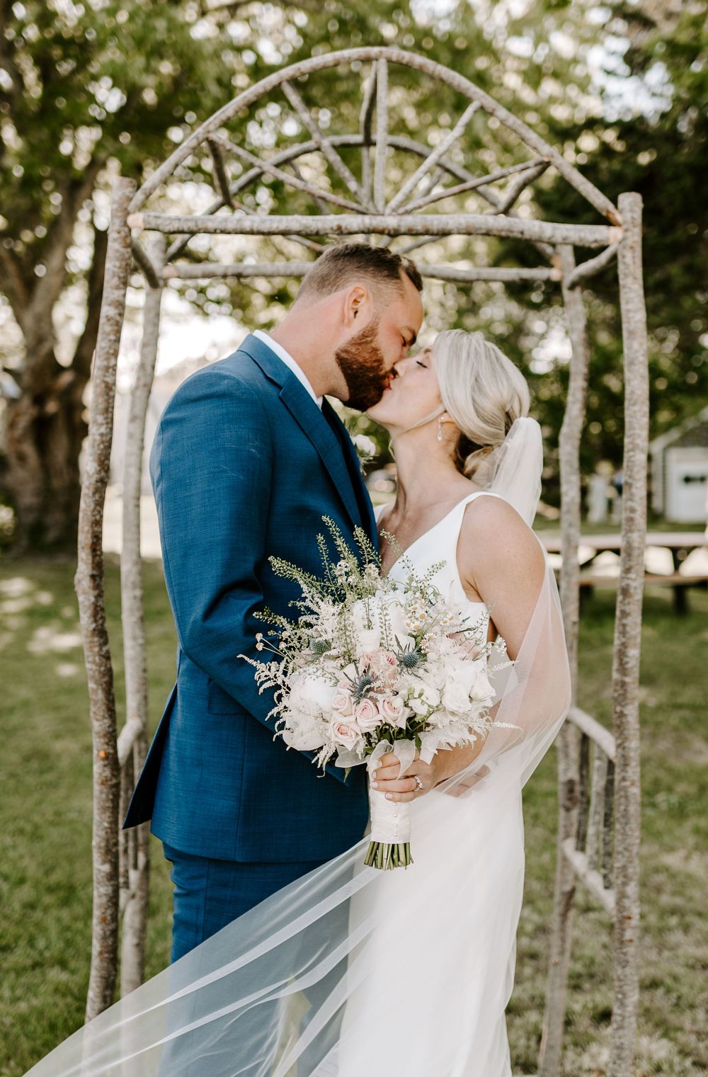 Romantic wedding kiss under rustic wooden arch with flowing veil.