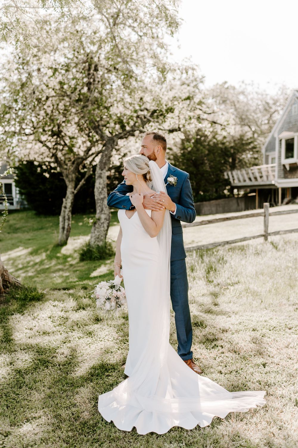 Sunlit wedding portrait showing a tender embrace between newlyweds on a grassy slope.