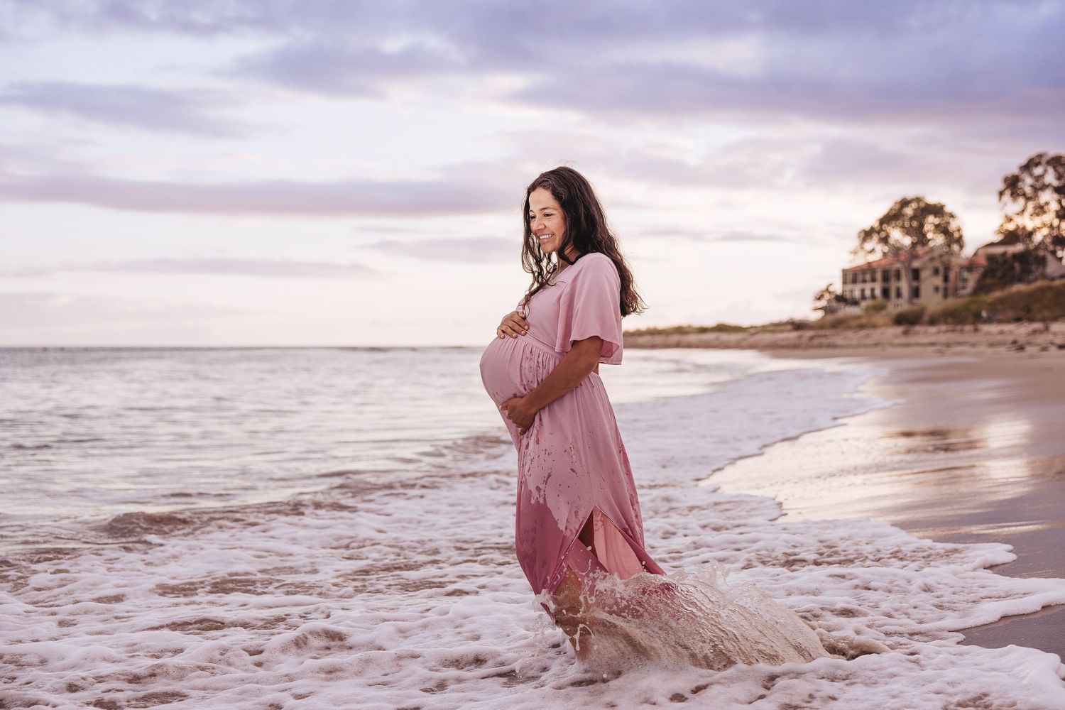 Woman in pink dress stands at sunset on a beach with waves lapping at her feet.