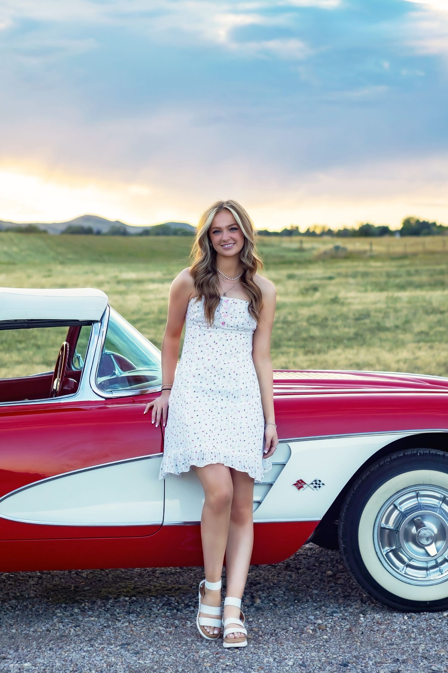 A girl poses with a classic red and white vintage car parked in field at sunset with mountains and golden sky in background.