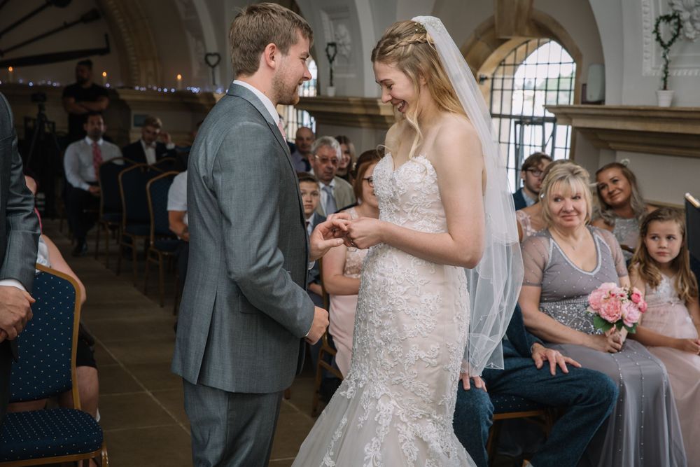 A couple exchanges rings during a wedding ceremony while guests watch from blue chairs in an ornate venue.
