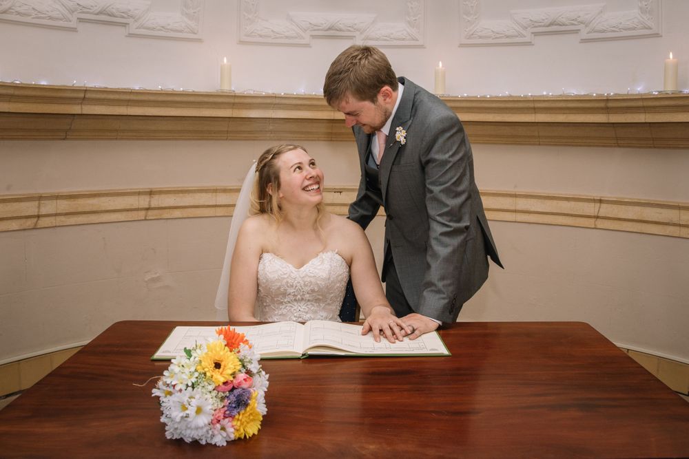 A wedding couple signs documents at a wooden table with a colorful bouquet.