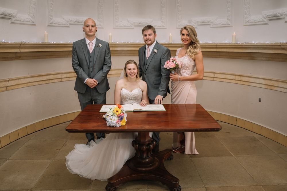 Wedding party gathers around an antique table during document signing in an elegant room.