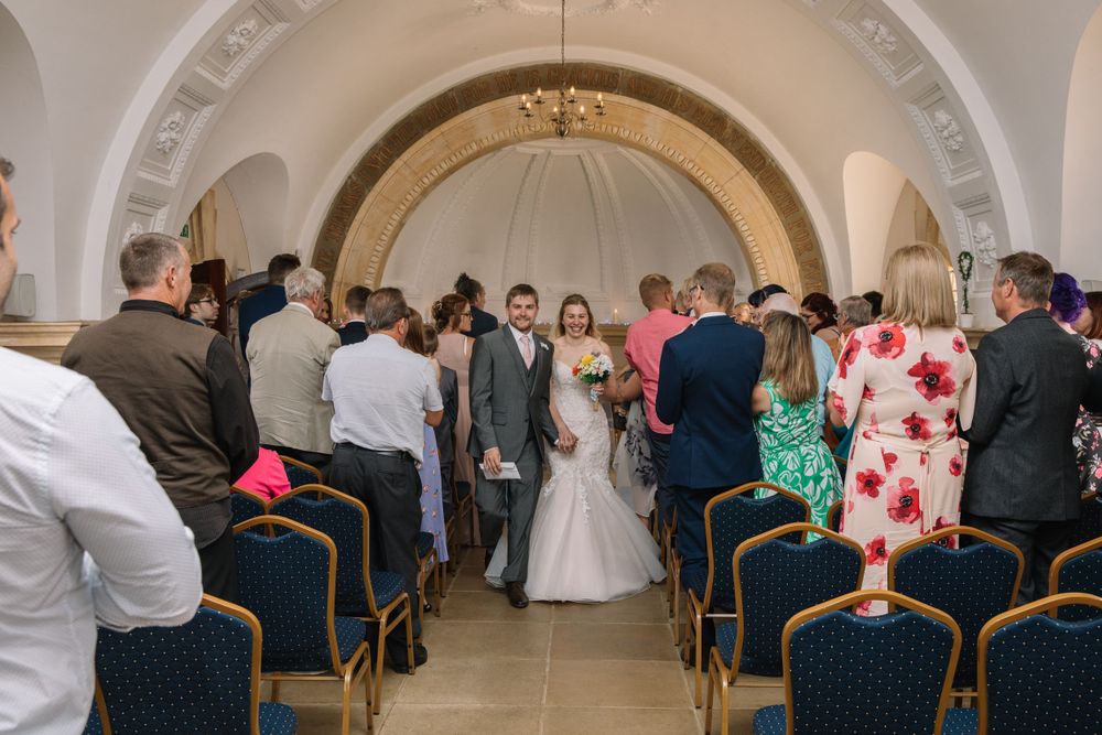 Wedding ceremony in progress under arched ceiling with guests seated in blue chairs.