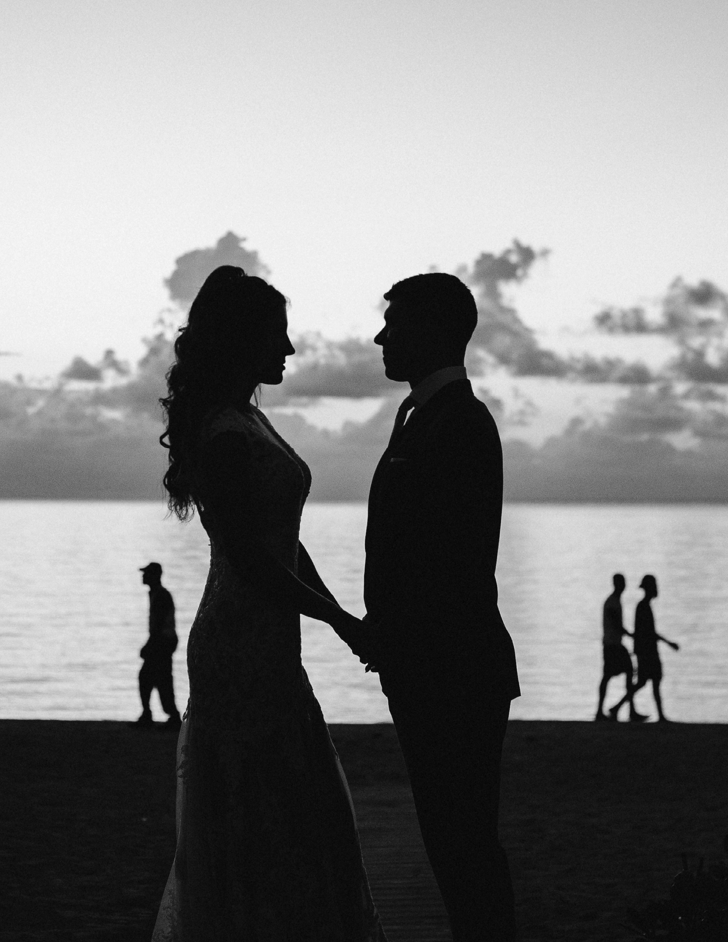 Silhouette of couple standing by ocean at sunset with people walking in background.