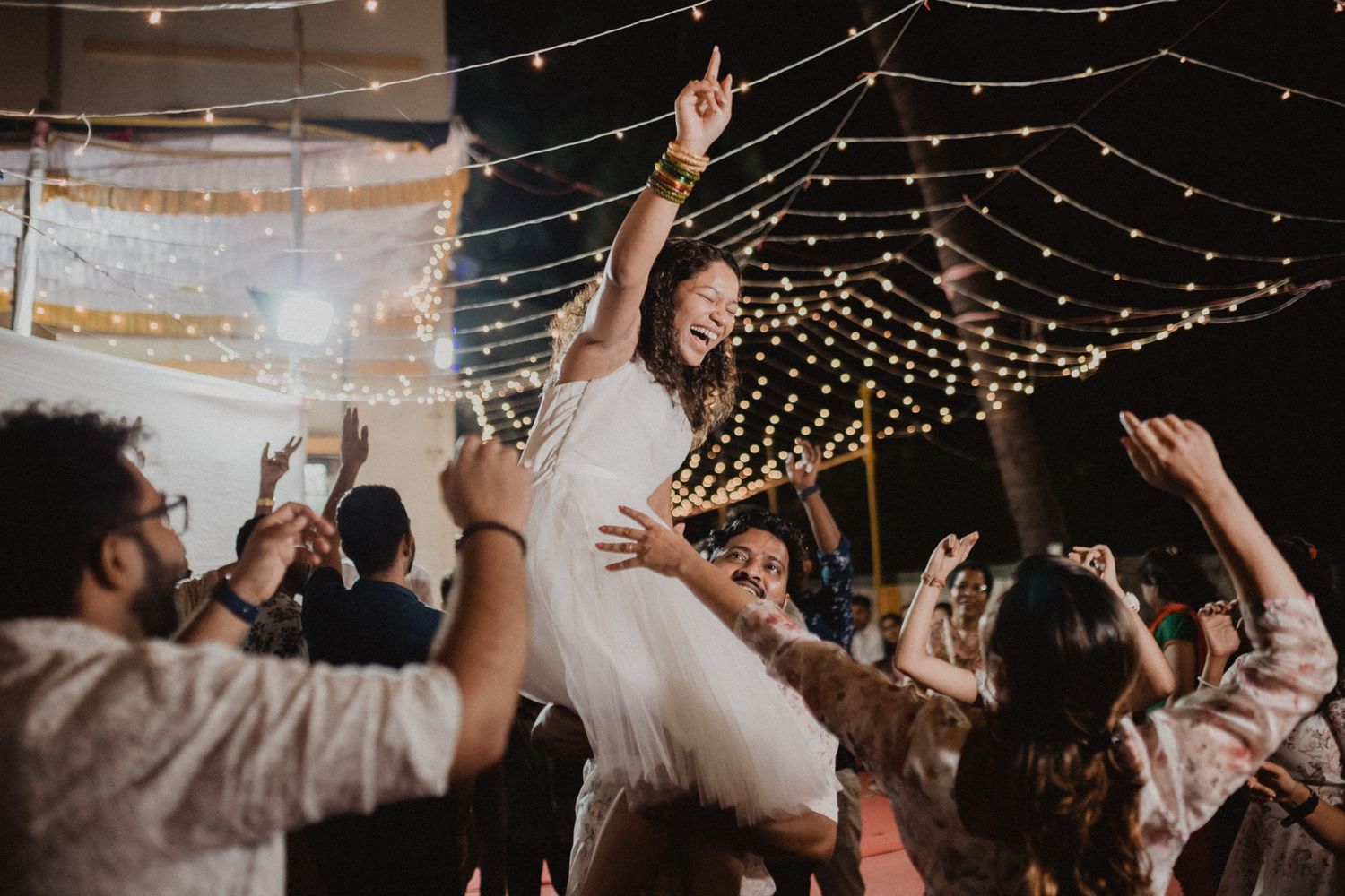 Party guests dancing and celebrating under romantic string light decorations in a dark venue.
