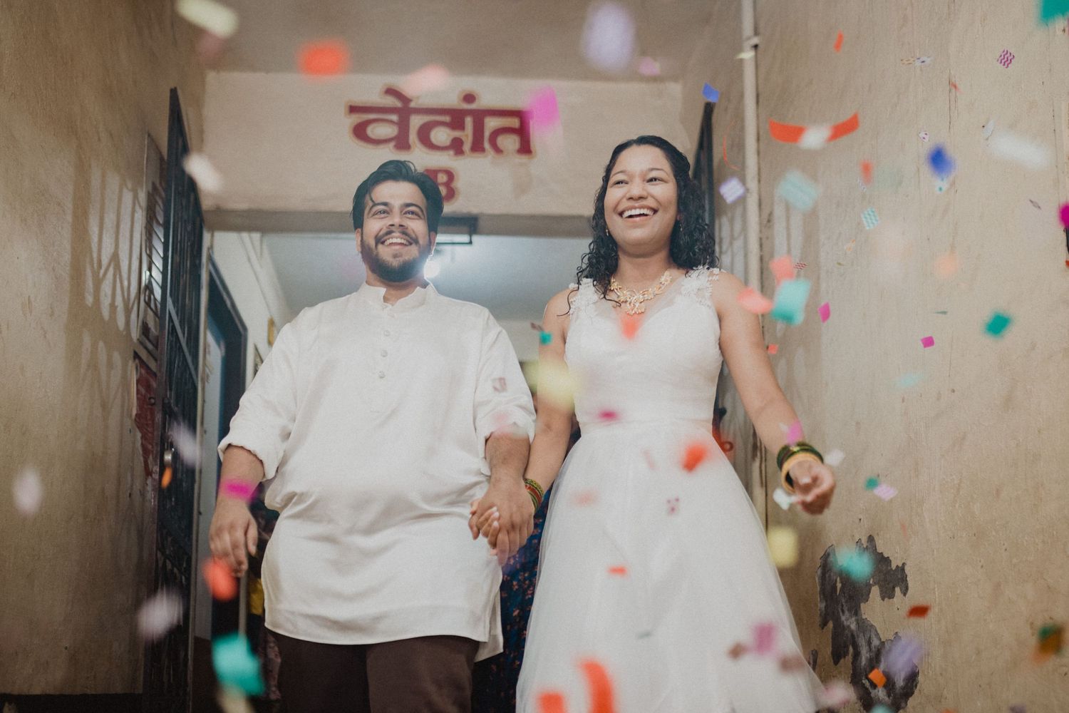 A couple joyfully walks through a hallway with confetti falling around them during a celebration.