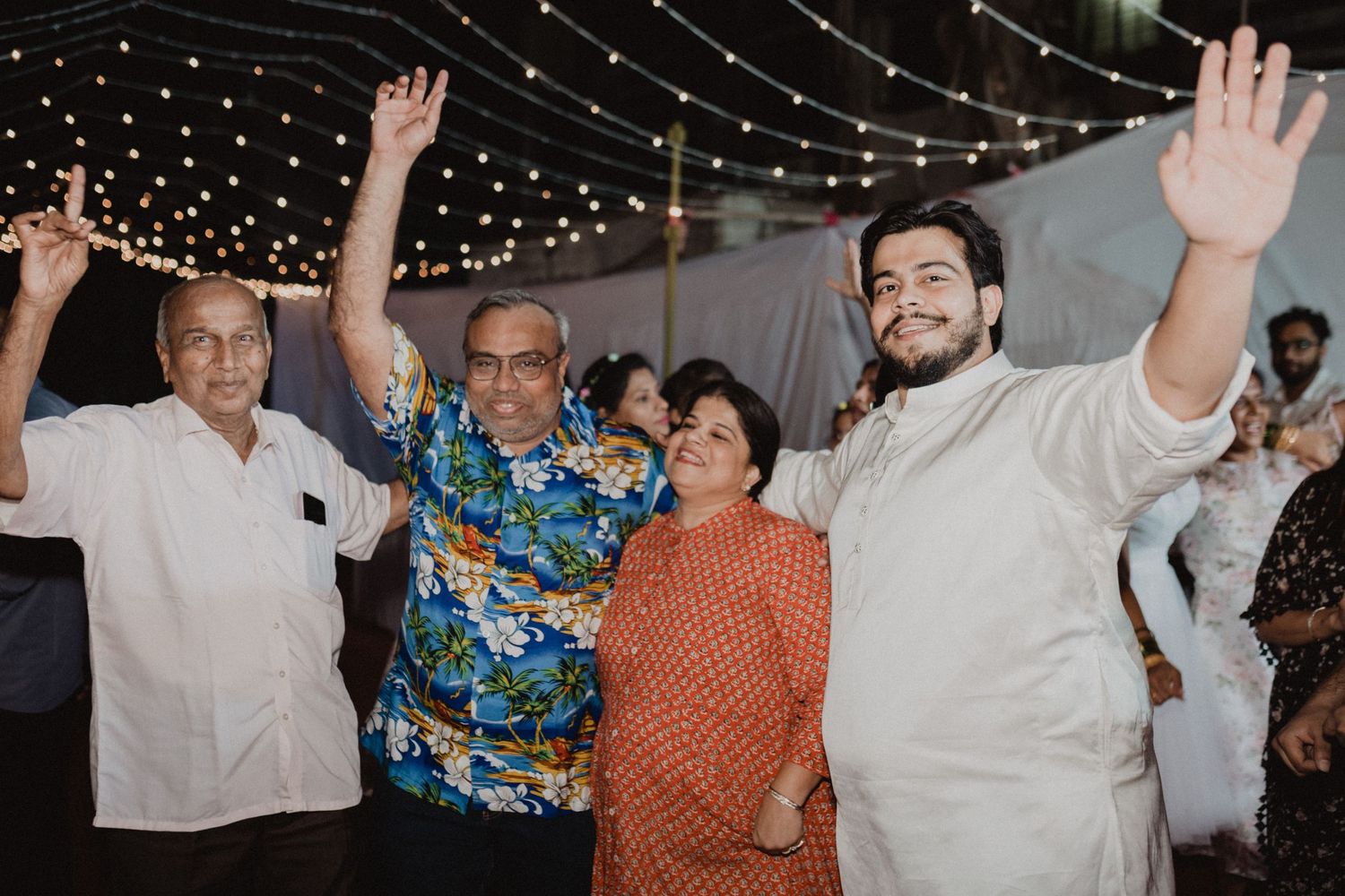 A group celebrates with raised hands under string lights at an outdoor evening event.