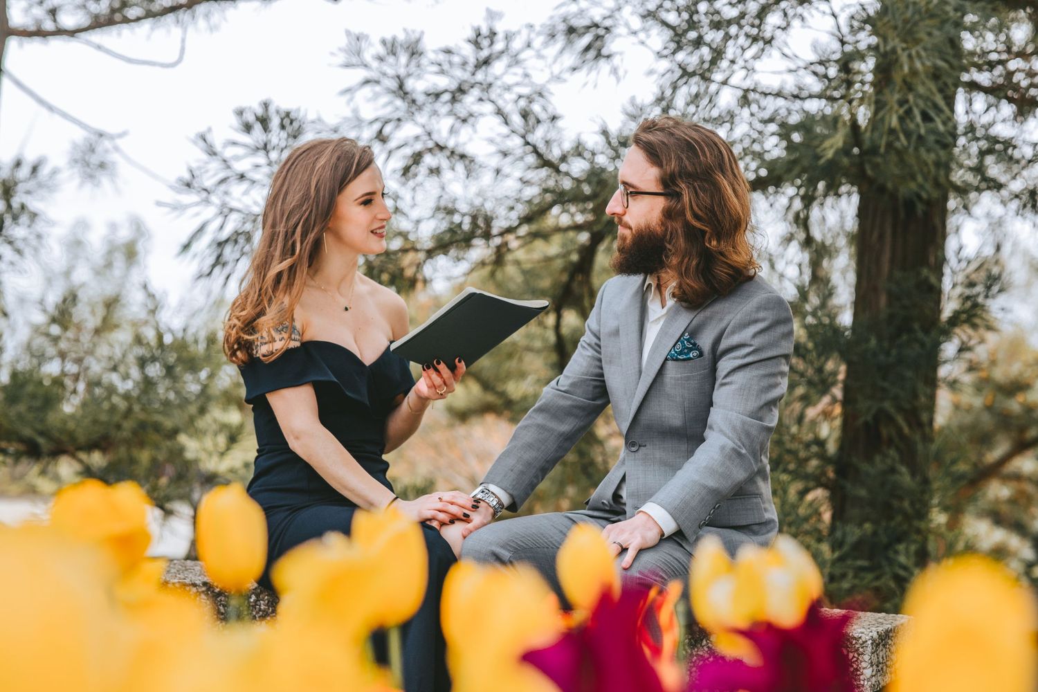 Couple sits together reading on bench surrounded by yellow tulips in garden setting.