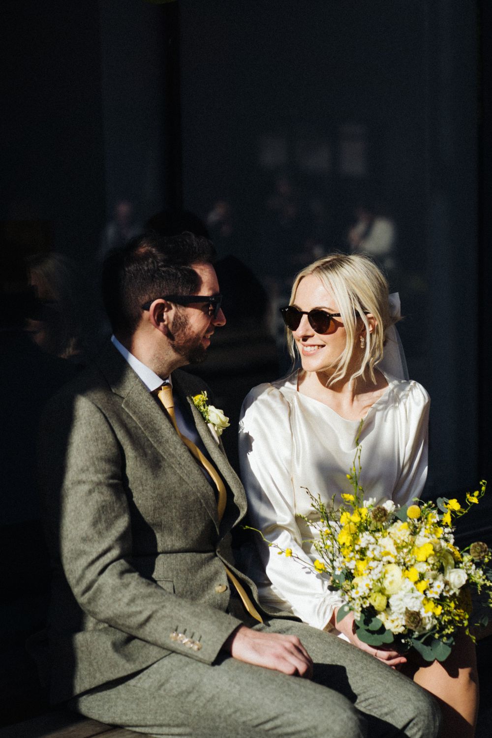 Wedding couple sharing a tender moment in sunlight with yellow bouquet against dark background.