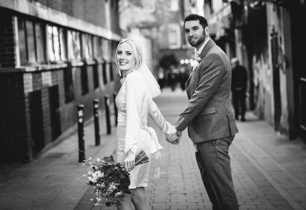 A black and white street portrait of a couple holding hands with urban architecture in background.