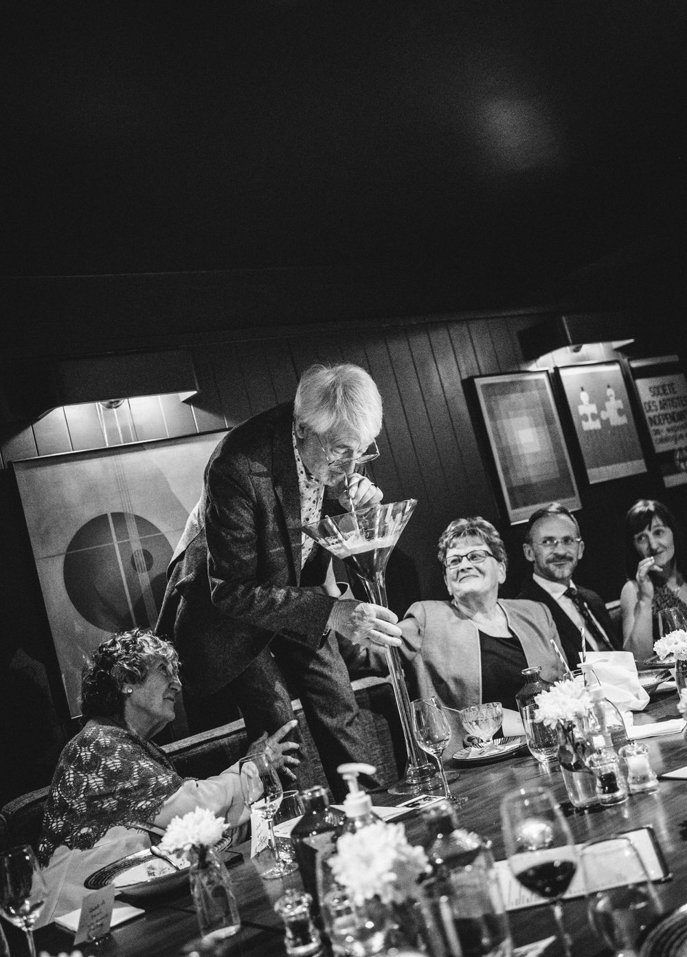 A black and white photograph of people enjoying drinks and conversation at a dinner table.