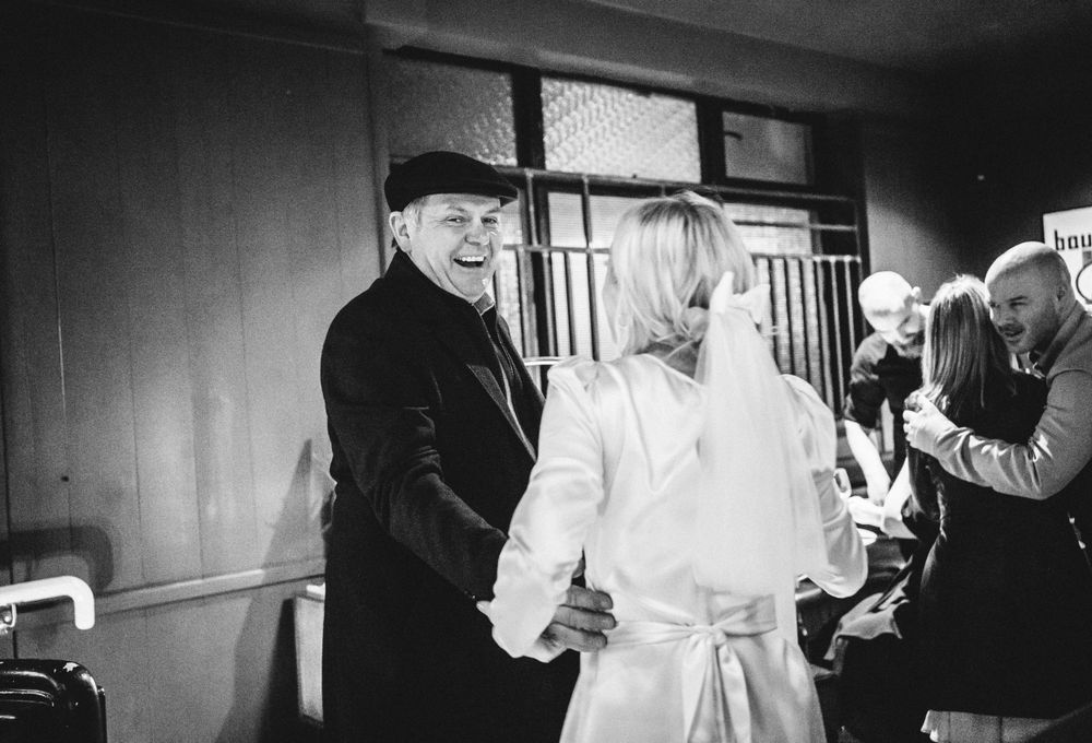 A black and white candid photo of wedding guests conversing at an indoor venue.
